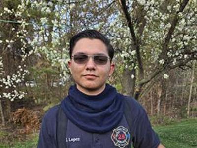 Jeydan Lopez, wearing a t-shirt from his fire department and holding a yellow helmet. He is standing in front of a cherry blossom tree.
