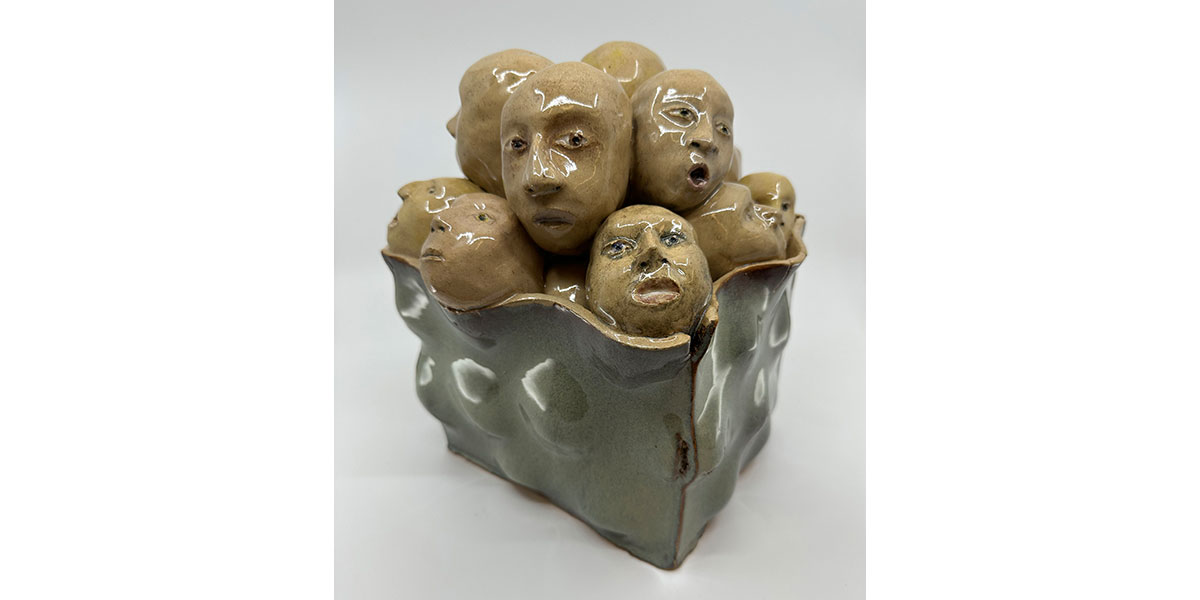 A glossy ceramic sculpture of a textured green vessel filled with a pile of tan-colored human heads with various facial expressions.