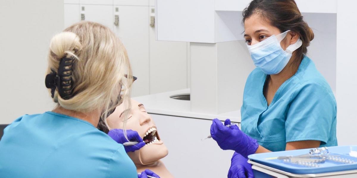 A dental assisting student in scrubs listens to instructions from her teacher.