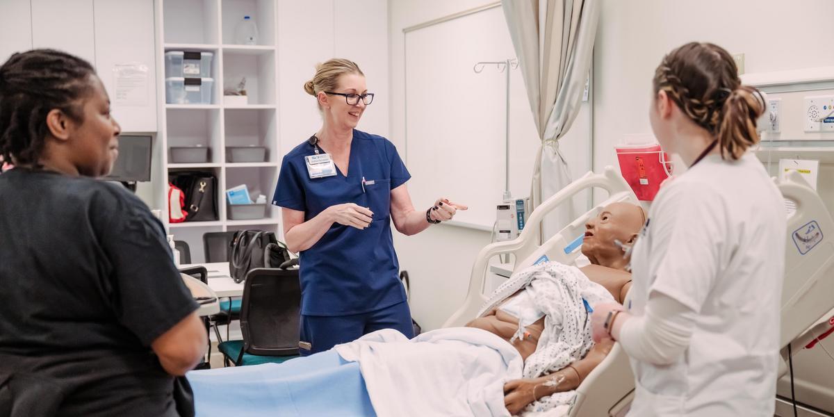 In a medical simulation lab, an instructor in blue scrubs guides two students around a medical mannequin lying in a hospital bed.